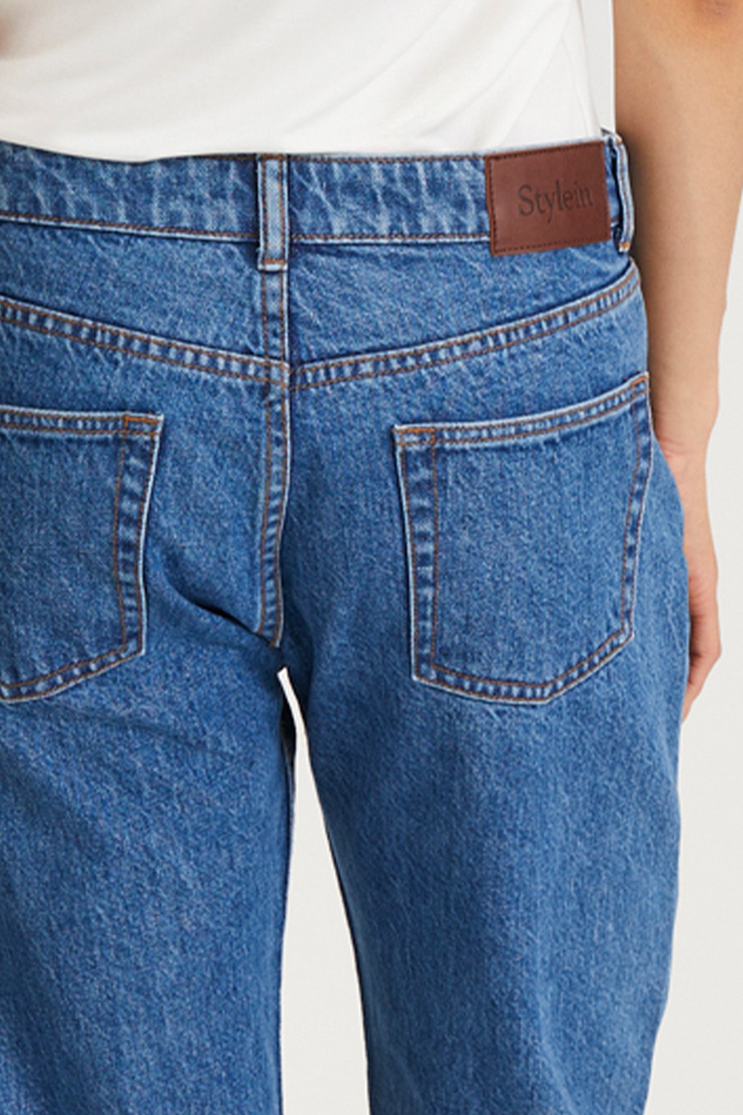 Close-up back view of a person wearing blue denim jeans with a white top, showing the waistband, belt loop, back pockets, and a brown leather patch with the brand name 'Stylein'.